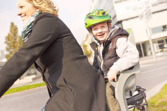 Eine blonde Frau fährt mit dem Fahrrad. Auf dem Kindersitz hinter ihr sitzt ein BUb. Er trägt einen Helm und lächelt.