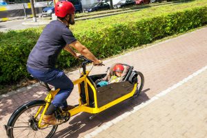 Ein Vater fährt mit einem gelben Lastenrad, im Korb vorne liegt seine 2 Jahre alte Tochter.