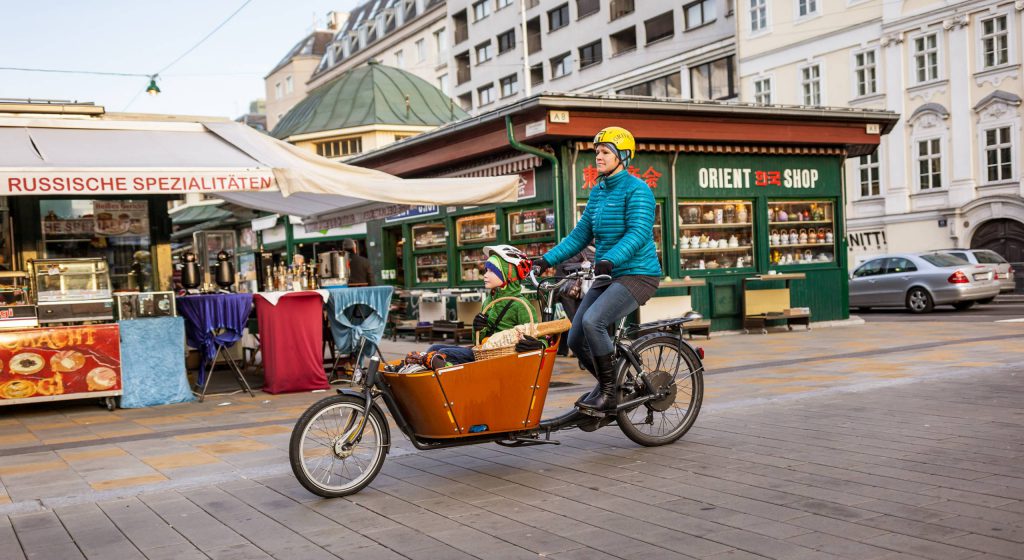 Frau fährt mit Transportfahrrad in der Begegnungszone Schleifmühlbrücke mit dem Naschmarkt im Hintergrund. Ihr kleiner Sohn sitzt im Korb des Transportfahrrads. Foto: Stephan Doleschal