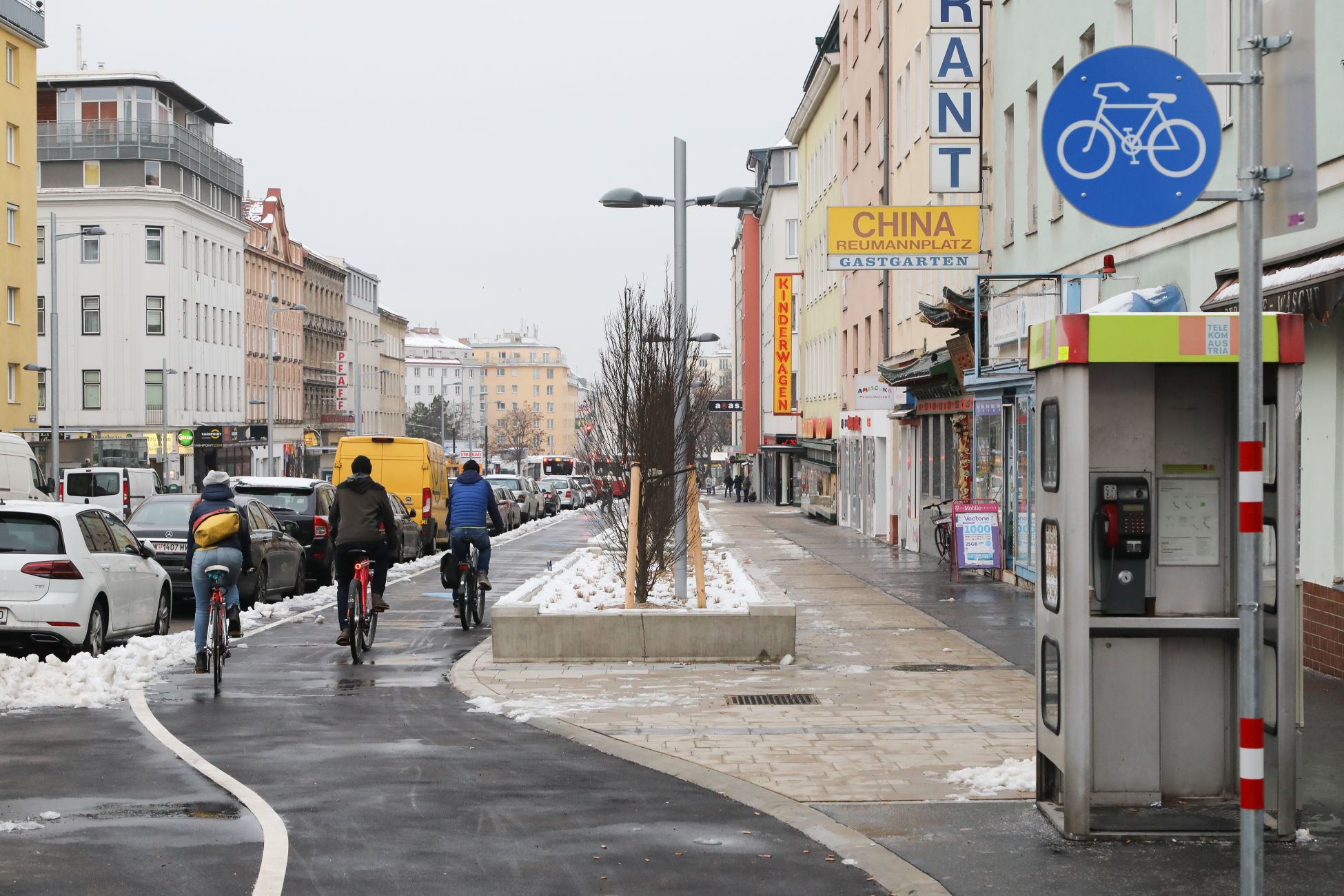 Drei Personen fahren auf dem Radweg entlang der Favoritenstraße, in Richtung Reumannplatz. Man sieht die Personen von hinten. Es ist Winter, am Straßenrand liegt Schnee. Foto aufgenommen von Christian Fürthner.