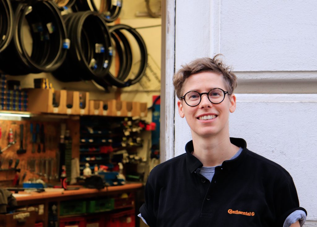 Pez steht vor der Fahrradwerkstatt, durch die offene Tür sieht man Reifen, die an der Wand hängen und verschiedene Werkzeuge auf einem langen Tisch