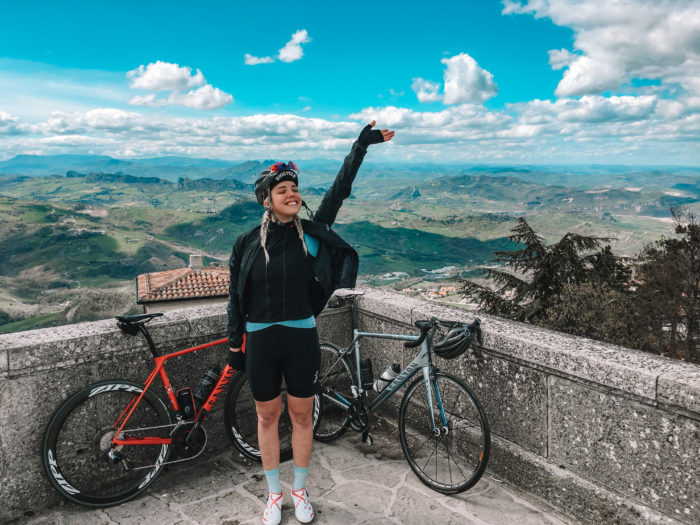 Nora steht mit zwei Fahrrädern auf einem Aussichtspunkt und streckt die Hand in die Höhe