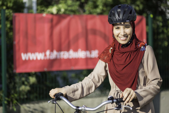 Eine Frau mit einem Schleier und Helm steht mit ihrem Fahrrad und lächelt in die Kamera